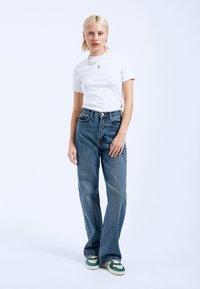 Witte t-shirt met korte mouwen, gecombineerd met blauwe wijde jeans en groen-witte sneakers. Model staat tegen een lichte achtergrond.