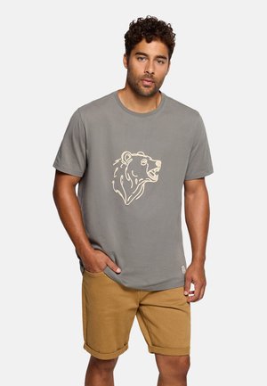 Hombre con camiseta gris con gráfico de cabeza de oso y pantalones cortos de color mostaza, de pie con una mano en el bolsillo contra un fondo liso.