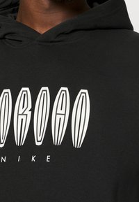Gros plan d'une personne portant un sweat à capuche noir avec de grandes lettres blanches stylisées et un texte plus petit indiquant Nike au niveau de la poitrine.