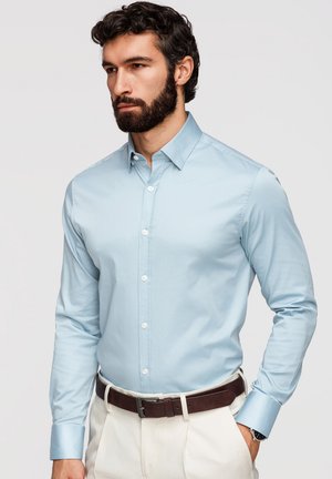 Homme barbu portant une chemise bleue claire à boutons et un pantalon blanc, ceinture marron, debout devant un fond uni.