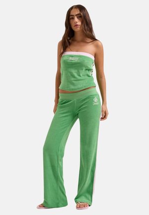 Jonge vrouw draagt een groene velours tube top en bijpassende wijde broek met "Juicy" logo, staand in een ontspannen houding op een witte achtergrond.