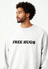 Sweat-shirt gris à manches raglan avec une impression noire "FREE HUGS". Le texte est en gras, légèrement en relief, sur un tissu doux et texturé.