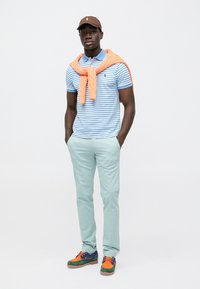 Polo shirt with blue and white stripes, orange sweater tied around the neck, mint green pants, and multicolored sneakers. Brown cap.