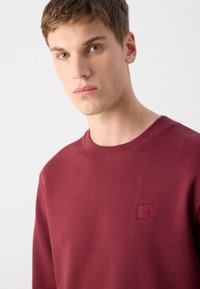 Sudadera burdeos con cuello redondo, textura suave y pequeño logotipo cuadrado bordado en el pecho.