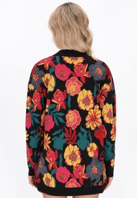 Donna con capelli biondi ondulati che indossa un maglione nero con un grande motivo floreale colorato in rosso, giallo e verde acqua, vista da dietro.