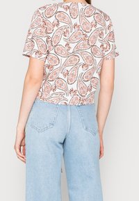 Donna che indossa una maglietta bianca a maniche corte con un motivo paisley rosso e jeans in denim chiaro a vita alta, vista da dietro.