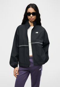 Γυναίκα που φοράει μαύρο μπουφάν με φερμουάρ και λευκό λογότυπο, λευκό κοντό τοπ, μοβ παντελόνι και μαύρα γυαλιά ηλίου, στέκεται μπροστά σε απλό φόντο.