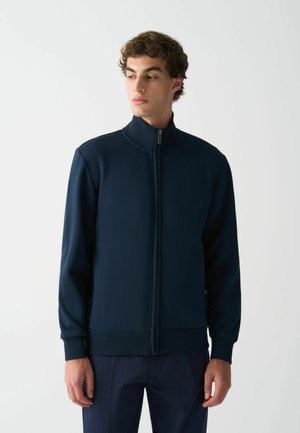 Marineblaue Zip-Jacke aus glattem Stoff mit Stehkragen, gerippten Bündchen und Saum. Verfügt über einen durchgehenden Reißverschluss und ein minimalistisches Design.