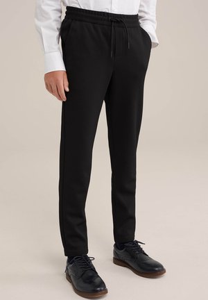 Mężczyzna w czarnych spodniach ze ściągaczem o fakturze, białej koszuli na guziki i czarnych skórzanych butach wizytowych stojący na jasnym tle.