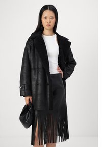Fekete műbőr kabát prémes gallérral, két elülső gombbal, fekete szoknyával párosítva, amely függőleges franekkel díszített.