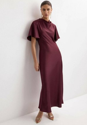 Vrouw die een bordeauxrode satijnen jurk tot op de vloer draagt met vlindermouwen, staand tegen een witte muur, en gouden open-toe hakken aan heeft.