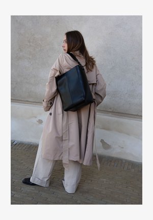 Femme en trench-coat beige et pantalon à jambes larges porte un grand sac cabas en cuir noir sur l'épaule devant un mur gris.