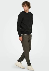 Schwarzer Strickpullover mit strukturiertem Muster, kombiniert mit grauen schmalen Hosen und weißen Turnschuhen, vor einem einfarbigen Hintergrund.