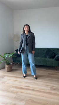 Blazer cinza ajustado sobre uma blusa preta, combinado com jeans claros de boca de sino e sapatos pretos de ponta. Sofá de veludo verde e lâmpada ao fundo.