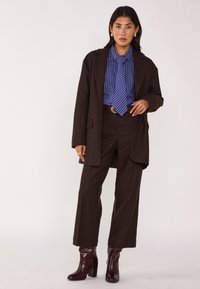 Abito marrone a righe con giacca larga e pantaloni a gamba larga, abbinato a una camicia blu a righe e una cravatta coordinata. Stivali bordeaux lucidi.