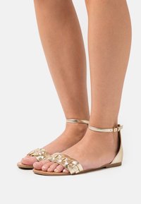 Gold flat sandals with woven straps, ankle buckle, and a smooth finish. Open-toe design and flat soles emphasize comfort and breathability.