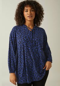 Blå leopardmönstrad topp med långa ärmar, rundad nederkant och en urgröpning vid halsen med knytdetalj. Mjuk tyglik med en slät yta.