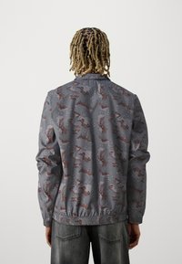 Grå camouflagejakke lavet af letvægtsmateriale, med elastiske manchetter og kant, prydet med brune og hvide accenter i mønsteret.