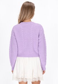 Ibolyakék kötött pulóver, fűzött mintával, lekerekített nyakkivágással és bordázott szegéllyel. Rövid fazon, fehér csipkés szoknyával párosítva.