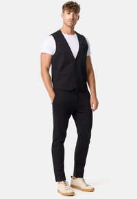 Schwarze Weste mit schmaler Passform und einreihigem Design, kombiniert mit schwarzen Hosen und beigen Sneakers. Darunter ein weißes T-Shirt.
