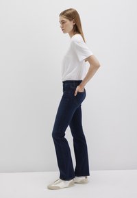 Flared dark blue jeans with a high waist, paired with a plain white T-shirt and white sneakers. Model stands sideways against a light background.