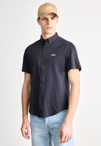 Tumehalli lühikeste varrukatega triiksärk kraega; rinnal valge BOSS-logo; koos kandes hele sinised teksapüksid ja beež nokamüts.