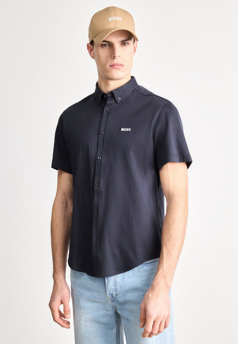 Tumehalli lühikeste varrukatega triiksärk kraega; rinnal valge BOSS-logo; koos kandes hele sinised teksapüksid ja beež nokamüts.