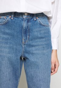 Osoba v modrých džínách s vysokým pasem a bílém dlouhém rukávu, s rukou uvolněně položenou vedle sebe.