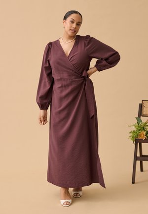 Femme portant une robe portefeuille à manches longues de couleur violet profond et des talons blancs ouverts aux orteils, debout avec une main sur la hanche devant un fond beige.