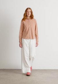 Long-sleeve, light peach shirt, button-up front, paired with loose white trousers and pink sneakers. Smooth fabric with a casual fit.