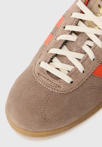 Bruna mockasin sneakers med vita snören, orangea sidremsor och en gummigummi sula, som visar detaljerad sömnad och textur.