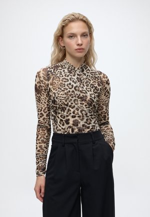 Femme aux longs cheveux blonds portant un haut transparent à manches longues imprimé léopard et un pantalon noir taille haute sur un fond uni.