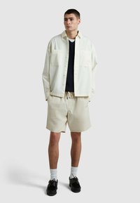 Giacca di camicia leggera color crema con due tasche frontali, indossata sopra un maglione scuro e pantaloni beige, completata da scarpe nere e calze bianche.