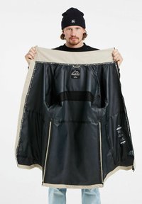 Giacca impermeabile beige e nera, con foderatura interna liscia, etichetta del logo e due tasche laterali. Il tessuto sembra resistente e leggero.