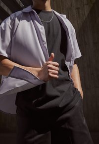 Camicia corta a maniche corte di colore viola chiaro con bottoni sopra una maglietta nera, abbinata a pantaloni scuri. Tessuto liscio, vestibilità rilassata, leggera lucentezza.