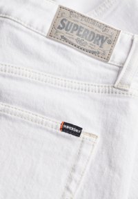 Denim blanc avec coutures brodées, portant une étiquette de marque grise et un petit tag noir Superdry avec un accent orange.