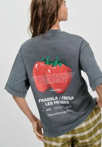 Camiseta gris de gran tamaño con un gran gráfico de fresa colorida en la espalda, con el texto "DELIGHT DE FRESA" en letras blancas y en negrita.