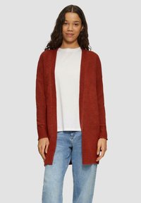 Cardigan tricoté couleur rouille, à devant ouvert, avec des manches longues et des poignets côtelés, superposé sur un t-shirt blanc et associé à un jean bleu.