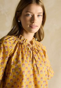 Kollane lilleline pluus, millel on kogutud kaelus, lühikesed varrukad ja lahtine lõige koos roosa ja rohelise lillemustriga.