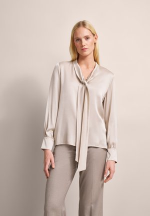 Beige Satinbluse mit langen Ärmeln und einer gebundenen Schleife am Kragen. Glatte Textur, lockere Passform und geraffte Manschetten. Kombiniert mit hellen Hosen.
