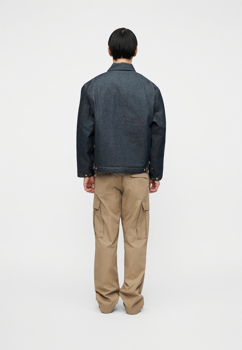 Veste en denim foncé à coupe classique, avec des boutons en accent et des coutures visibles. Associée à un pantalon cargo ample couleur kaki clair.