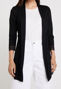 Svart öppen cardigan med trekvarts ärmar i spets, parat med en vit topp och vita högmidjade byxor, mjukt tyg och draperad design.