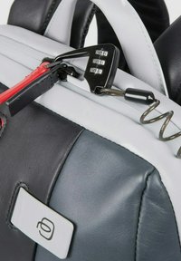 Zaino testurizzato nero e grigio con accenti bianchi, dotato di una serratura metallica per cerniera, un tirante rosso e un'etichetta con logo distintivo.