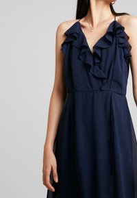 Robe bleu marine avec un col à volants, une découpe en V et des bretelles fines. Confectionnée dans un tissu léger avec une silhouette fluide.