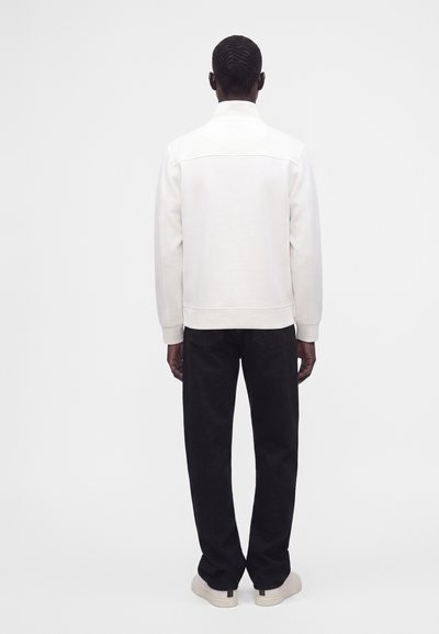 Homme de dos portant une veste blanche, un pantalon noir et des chaussures claires sur un fond blanc uni.