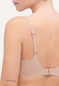 Nude wireless bra with adjustable straps, textured stretch fabric, and hook closure. Features a subtle logo on the strap.