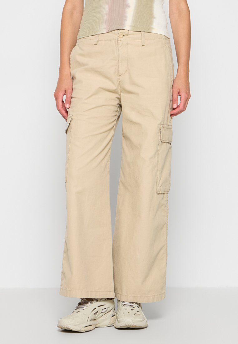 Pantalones cargo beige de pierna ancha, hechos de una mezcla de algodón. Cuentan con bolsillos laterales, un cierre de botón y una tela suave y texturizada.