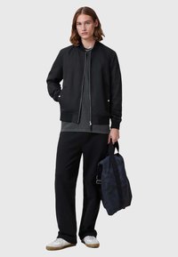Schwarze Bomberjacke, graues T-Shirt darunter, lockere schwarze Hose, weiße Sneakers, hält eine Tasche in dunklem Camouflage; glattes Material, lässiges Design.