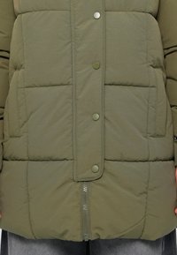 Maslinasto zeleni puffer kaput s kvadriranim uzorkom, dugmadi na pritisak i prednjim zipom, s dva bočna džepa. Izrađeno od izdržljive tkanine.