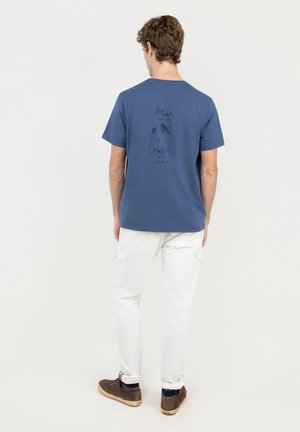 Hombre con camiseta gráfica azul, pantalones cargo blancos y zapatos marrones, de pie con la espalda hacia nosotros contra un fondo sencillo y claro.